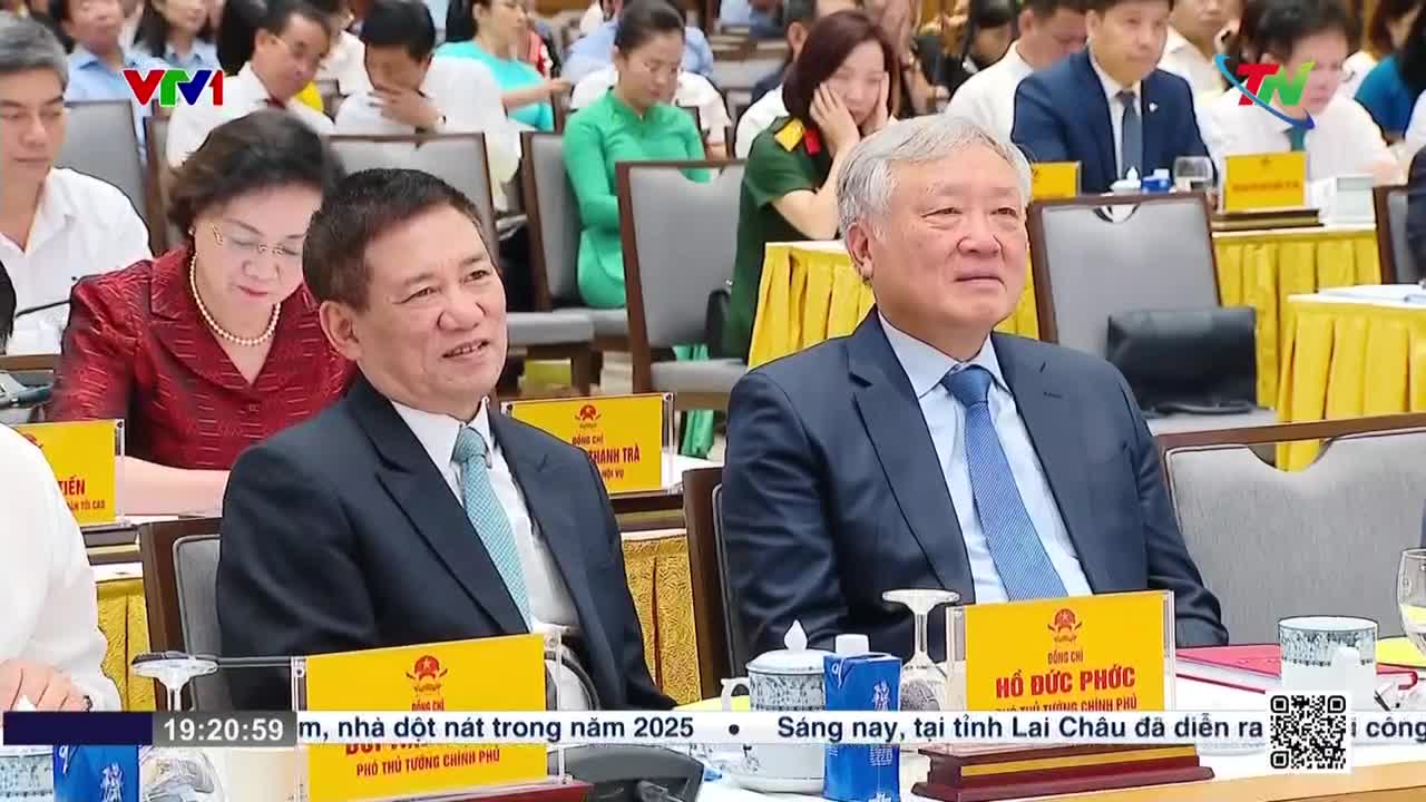 Two men in suits sit at a table, listening intently as the Thai Nguyen TV broadcast rolls across the bottom of the screen. The man on the right, wearing a blue tie, seems to be the focus of the camera.
