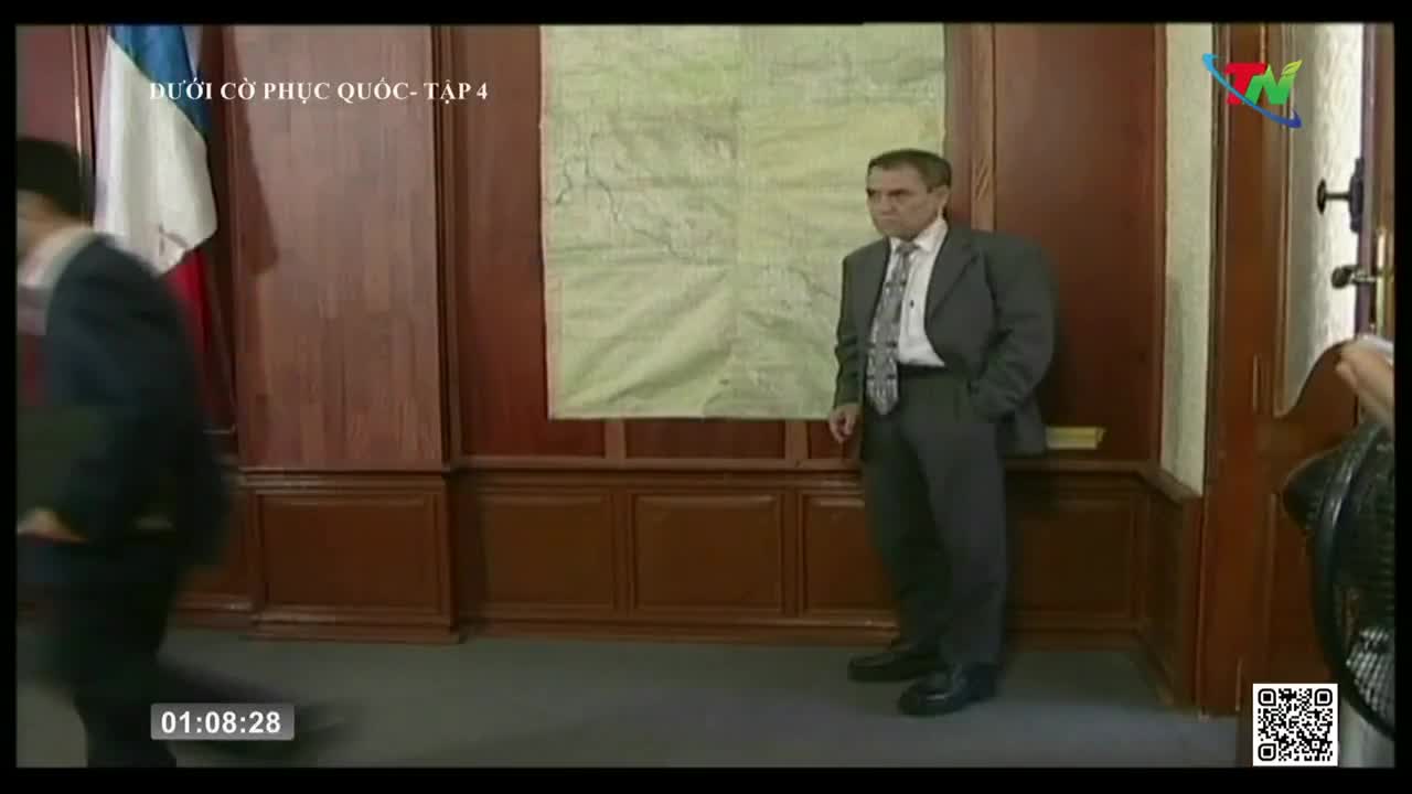 A man in a suit stands near a large map, his hands in his pockets, as another figure quickly walks past. The scene is being broadcast on Thai Nguyen TV in Vietnam.
