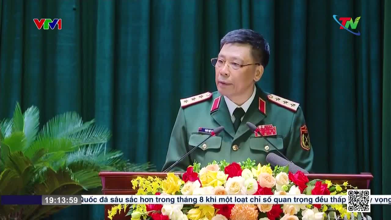 A man in a military uniform speaks from behind a podium decorated with flowers, as the Vietnam Television logo is visible in the upper left corner. The Thai Nguyen TV logo is in the upper right, and text scrolls at the bottom of the screen.
