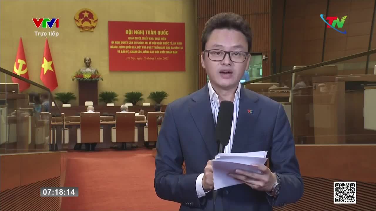 A man in a suit and glasses speaks into a microphone, holding papers in his hands, likely reporting live for Vietnam Television. Behind him, a formal meeting is underway, with flags and a podium visible.
