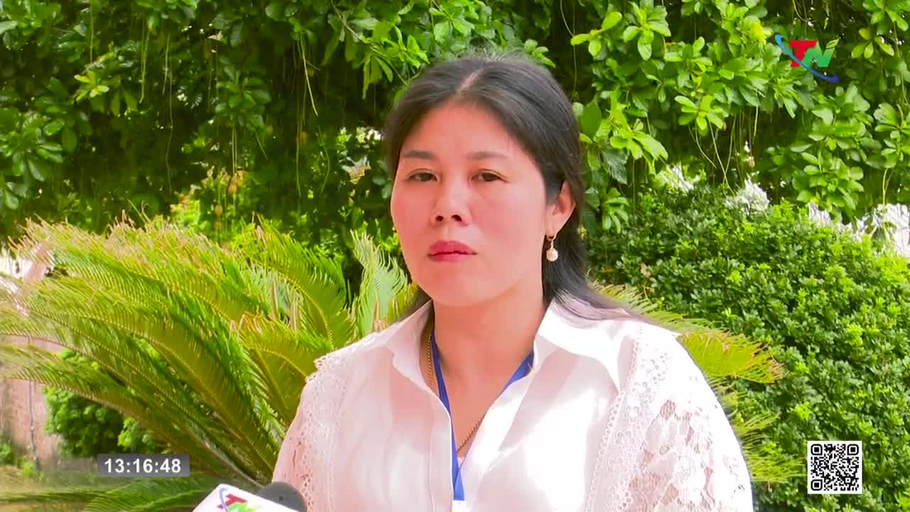 A woman in a white blouse speaks directly to the camera, the Thai Nguyen TV logo visible in the corner. Lush green foliage forms a backdrop as she delivers her message.
