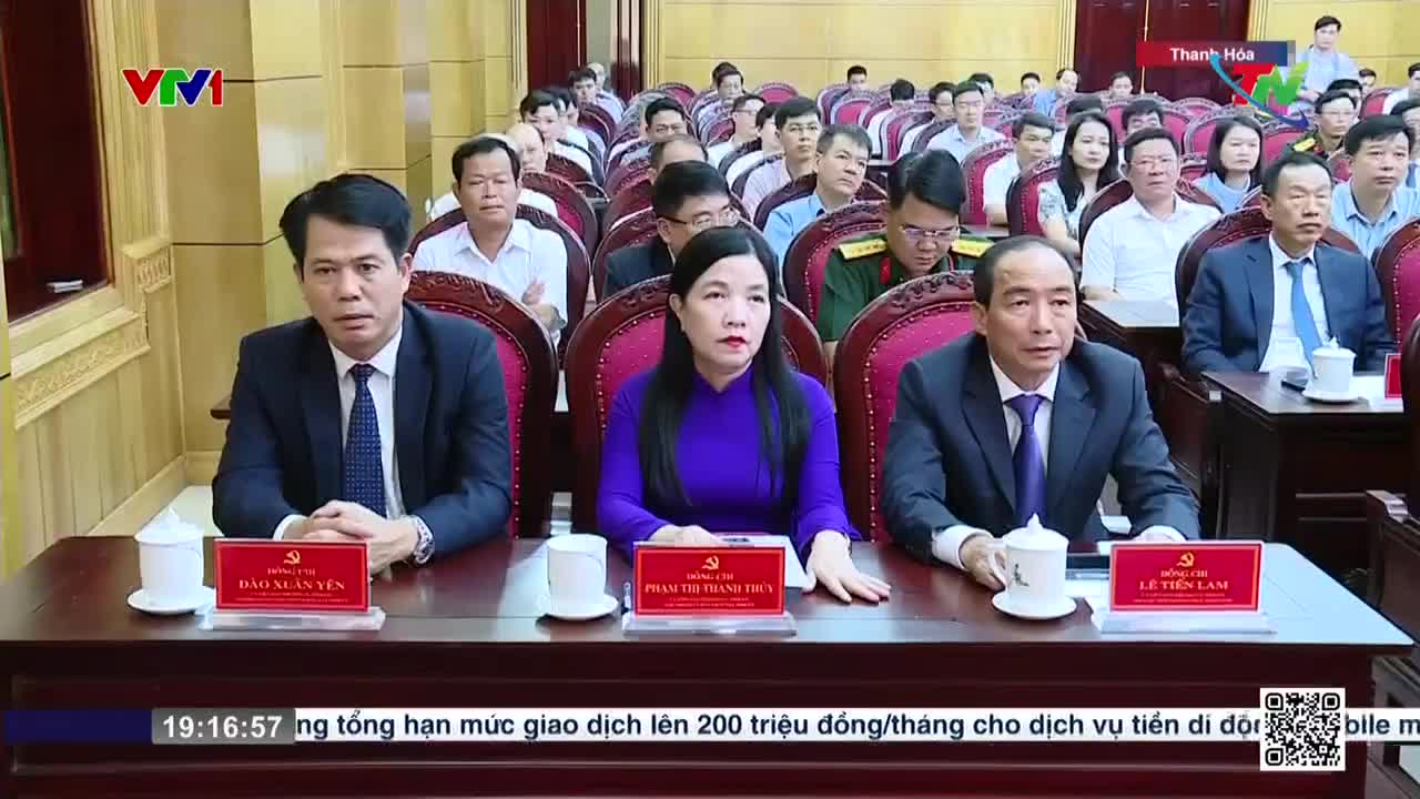 In a wood-paneled room, three officials sit at a long table, each with a nameplate in front of them. Behind them, a crowd of people in formal attire fill the rows of chairs, all focused on the proceedings, which are being broadcast by VTV1.
