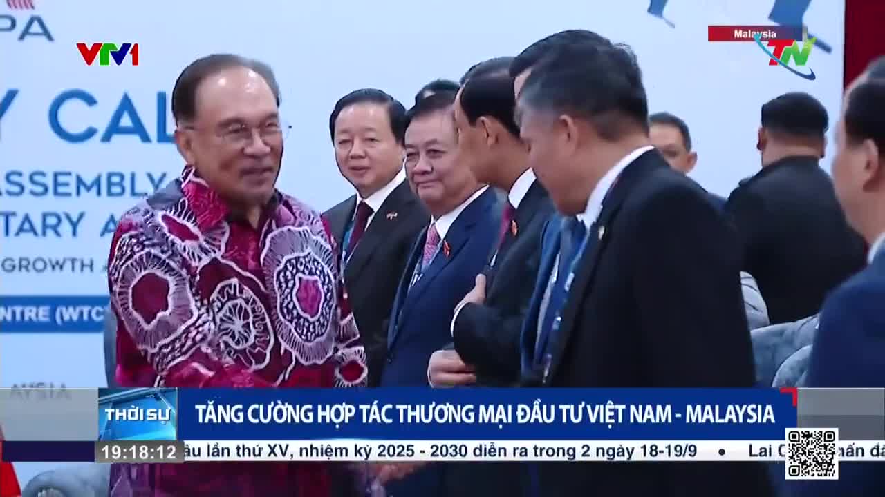 A man in a patterned shirt smiles as he chats with a group of men in suits. The scene is broadcast on Vietnam's VTV1 and Thai Nguyen TV, with a headline about Vietnam-Malaysia trade cooperation.
