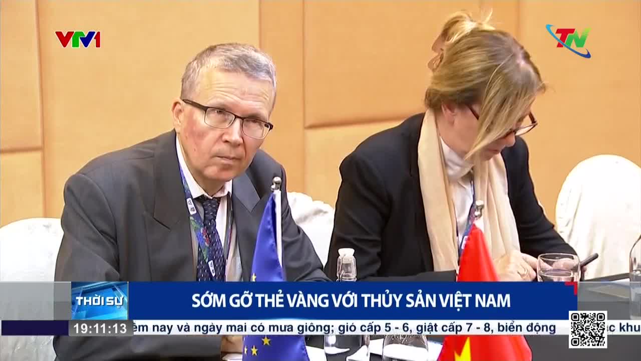 Two people, both wearing glasses, sit at a table with small flags in front of them. The man on the left looks directly at the camera, while the woman on the right is looking down, with a scarf around her neck.
