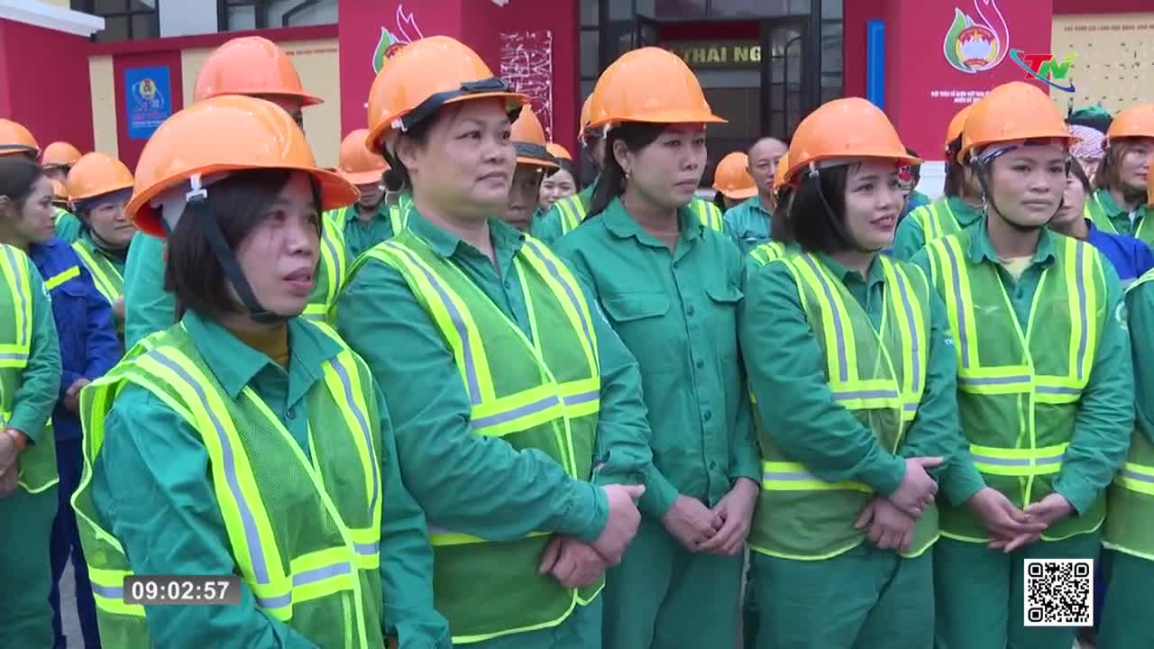 A line of women in green uniforms and orange hard hats stand ready. Their bright yellow safety vests catch the light as they face forward.