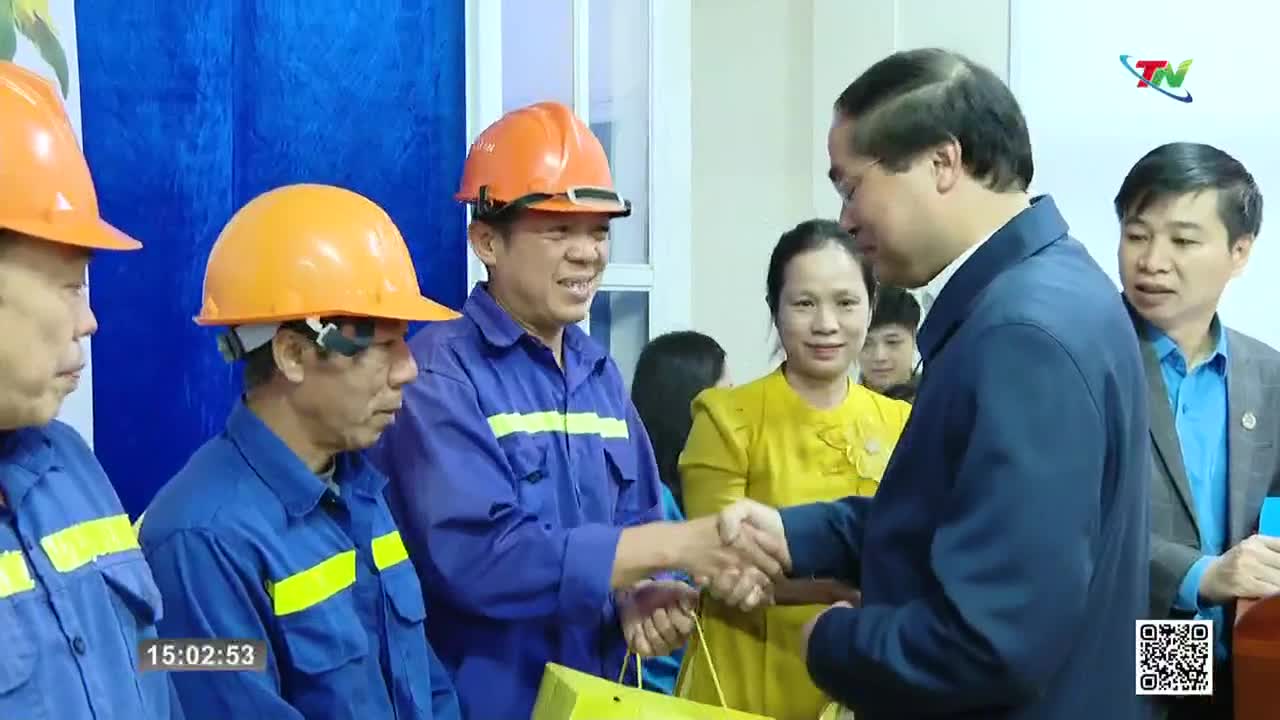 A man in a blue suit shakes hands with a worker wearing an orange hard hat and blue overalls. Another worker, also in an orange hard hat, smiles as he watches the exchange.