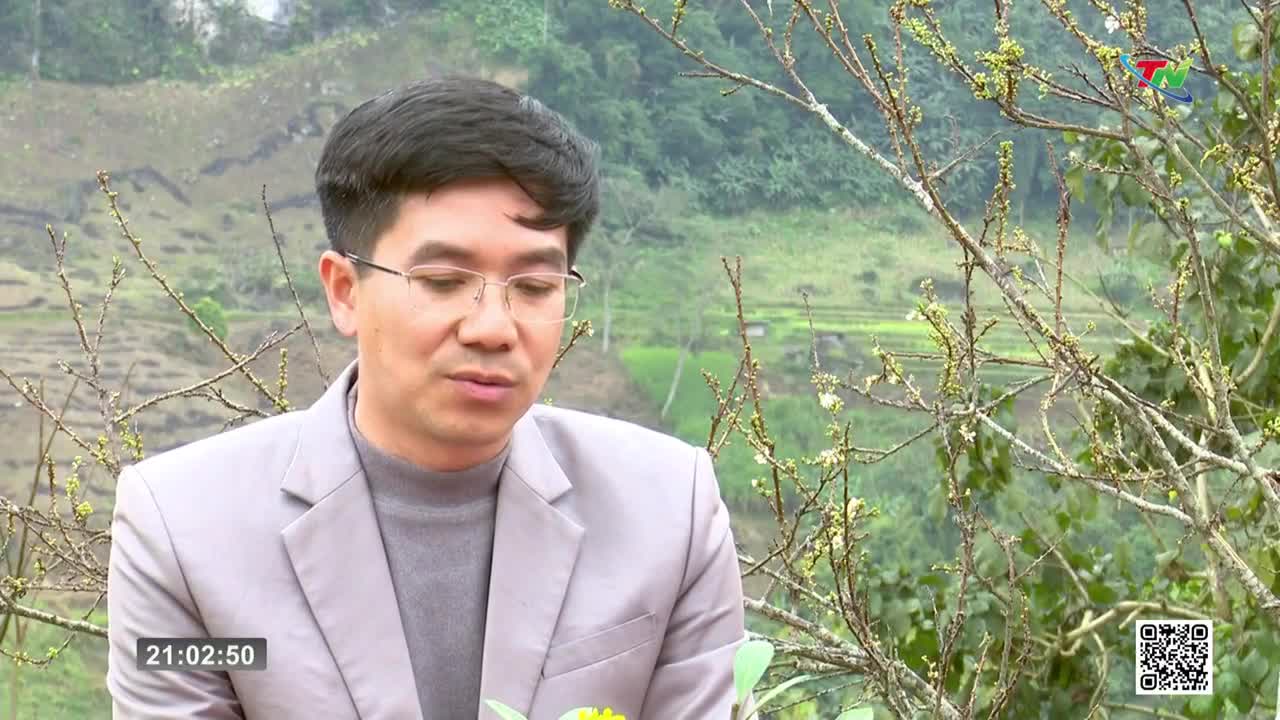 A man in a light suit and glasses speaks, holding a yellow flower. Behind him, the green terraces of Vietnam stretch into the distance.