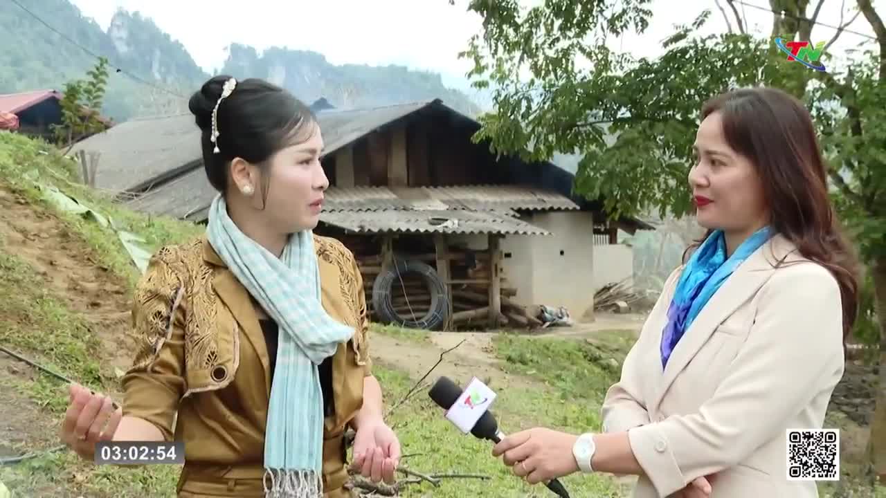 A woman in a patterned jacket and light blue scarf speaks into a microphone held by another woman. Behind them, a rustic house stands against a backdrop of green hills.