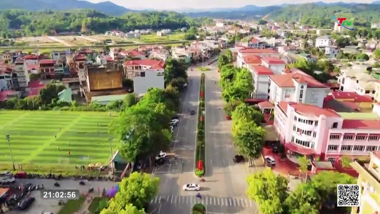 A white car cruises down the wide, tree-lined avenue in Thai Nguyen. Across the street, players are scattered across a bright green soccer field.