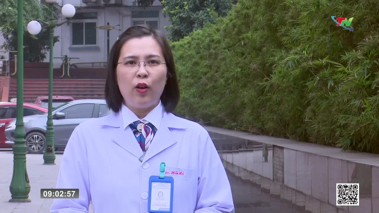 A woman in a white lab coat stands before a wall of bamboo, speaking into a microphone. Cars are parked behind her, and a timestamp reads 09:02:57.