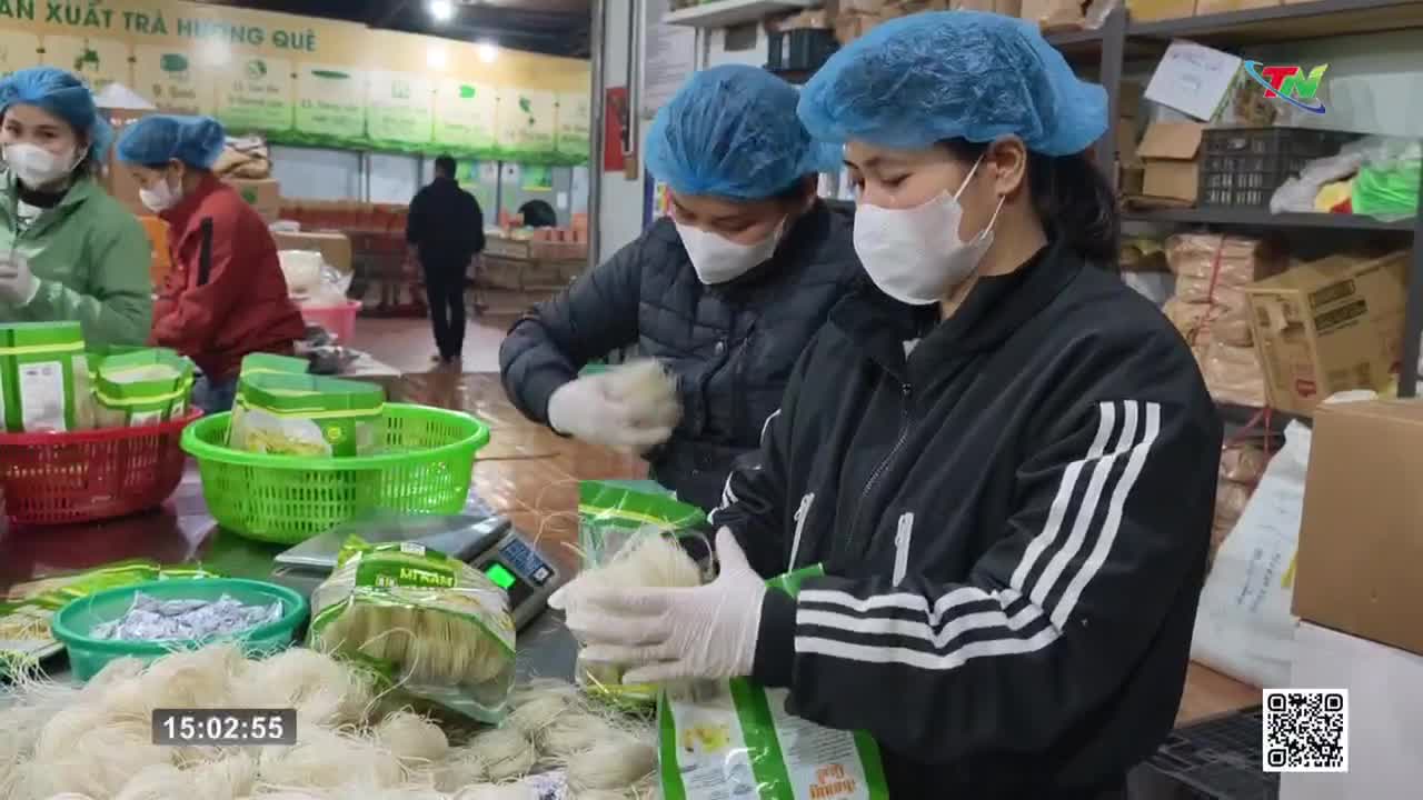 Workers in blue hairnets and masks are packaging dried noodles. A digital scale sits on the table beside them.