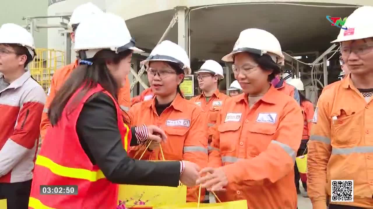 A woman in a red vest hands a yellow gift bag to another woman wearing an orange jumpsuit. Several other workers in similar attire stand nearby, their faces turned towards the exchange.