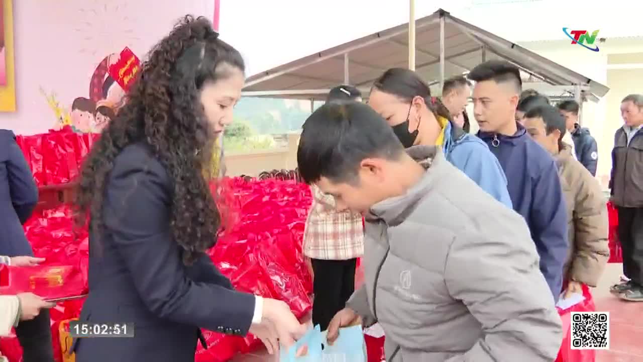 A woman in a dark suit hands a blue bag to a man in a gray jacket, while others wait in line behind him. The scene unfolds outdoors, with a backdrop of red bags and a banner featuring cartoon figures.
