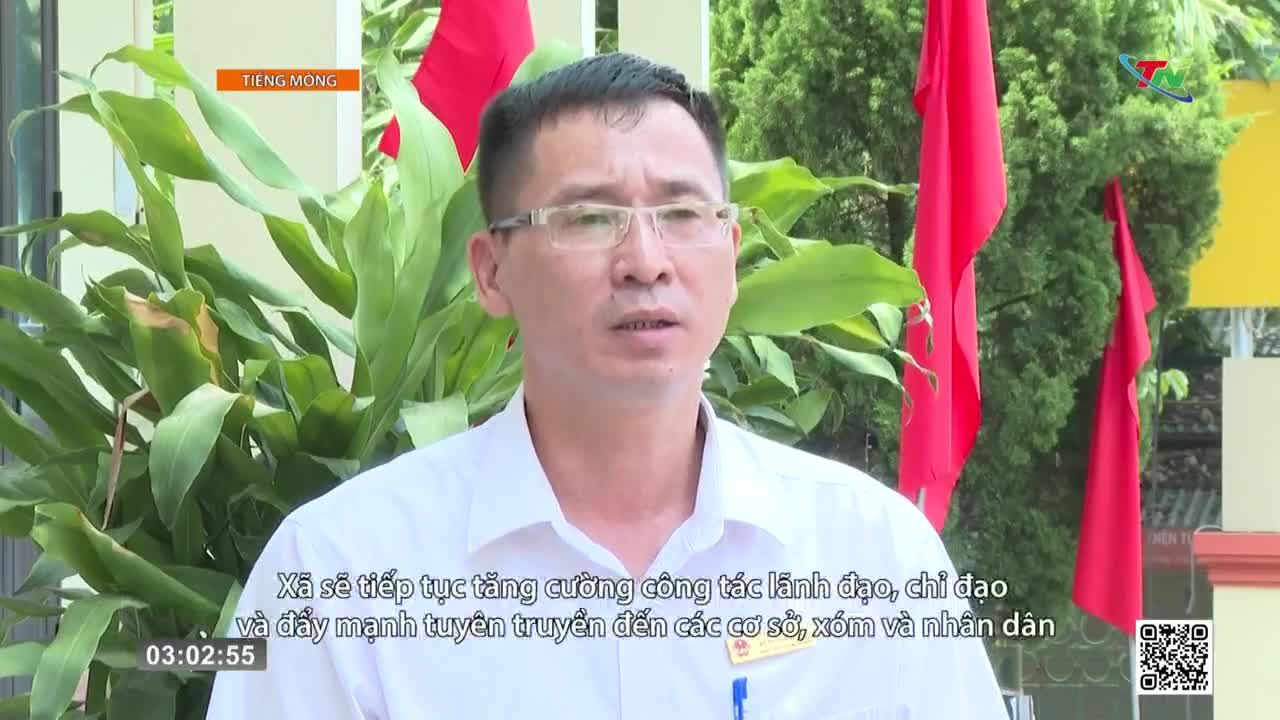 A man in a white shirt speaks, his glasses reflecting the light. Red flags hang behind him, their fabric still in the breeze.
