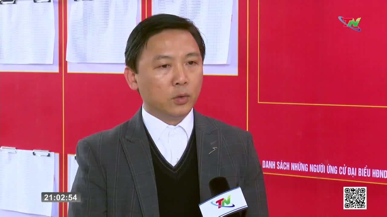 A man in a suit speaks into a microphone, his words being broadcast by Thai Nguyen TV. Behind him, lists of names are posted on a bright red wall.