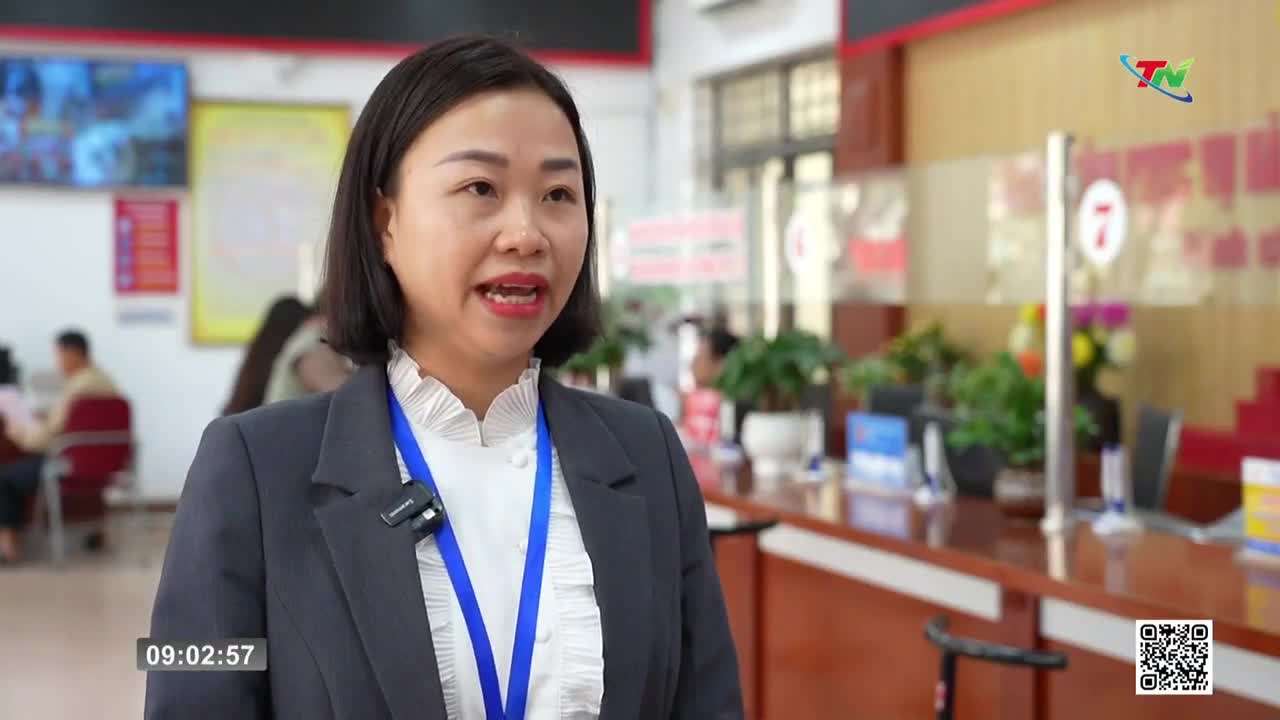 A woman in a dark suit speaks into a microphone, her white ruffled collar visible. Behind her, a busy office setting with people at desks and potted plants suggests a government office in Thai Nguyen. A woman in a dark suit speaks into a microphone, her white ruffled collar visible. Behind her, a busy office setting with people at desks and potted plants suggests a government office in Thai Nguyen.