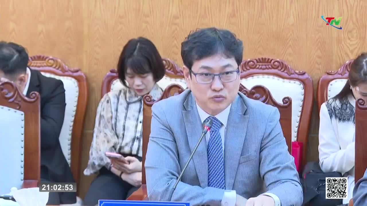 A man in a gray suit speaks into a microphone, his glasses reflecting the light. To his left, a woman in a patterned blouse looks down at her phone. A man in a gray suit speaks into a microphone, his glasses reflecting the light. To his left, a woman in a patterned blouse looks down at her phone.