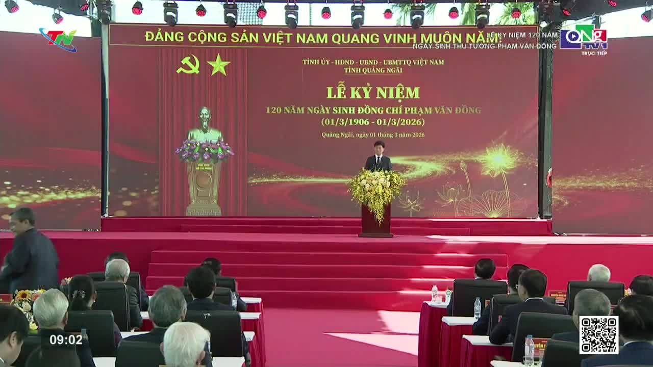 A man stands at a podium, addressing an audience seated on a red carpeted stage. Behind him, a large screen displays Vietnamese text and a golden bust. A man stands at a podium, addressing an audience seated on a red carpeted stage. Behind him, a large screen displays Vietnamese text and a golden bust.