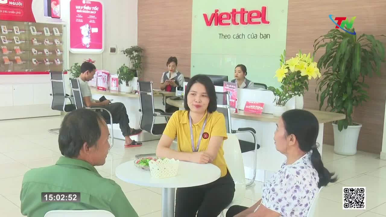 A woman in a yellow shirt speaks with a customer at a small table. Behind them, other customers wait while staff members assist them at counters. The Viettel logo hangs prominently on the wall. A woman in a yellow shirt speaks with a customer at a small table. Behind them, other customers wait while staff members assist them at counters. The Viettel logo hangs prominently on the wall.