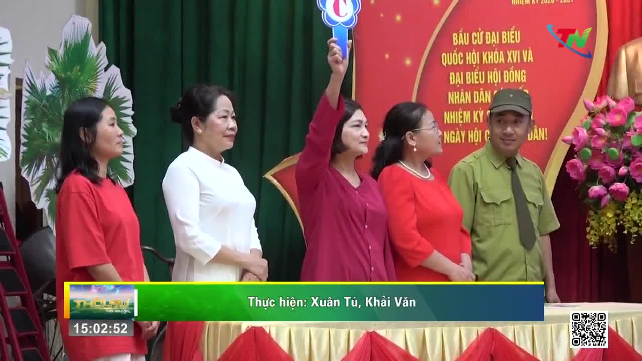A woman in a magenta top raises a blue paddle with a white symbol. Beside her, a man in a green uniform and cap smiles. A woman in a magenta top raises a blue paddle with a white symbol. Beside her, a man in a green uniform and cap smiles.