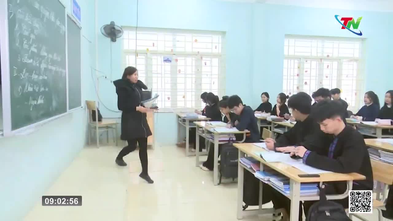A teacher in a black coat walks across the front of the classroom, holding papers. Students bent over their desks, writing.