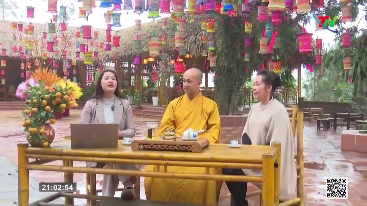 A woman in a tan blazer speaks, facing a man in a saffron robe and another woman in a cream shawl. Colorful lanterns hang from trees above a bamboo table set with tea.