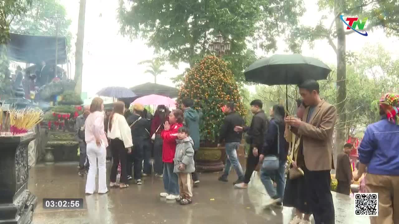 A group of people gather under umbrellas as incense smoke rises from a shrine. A man in a brown jacket checks his phone while holding an umbrella over a woman.