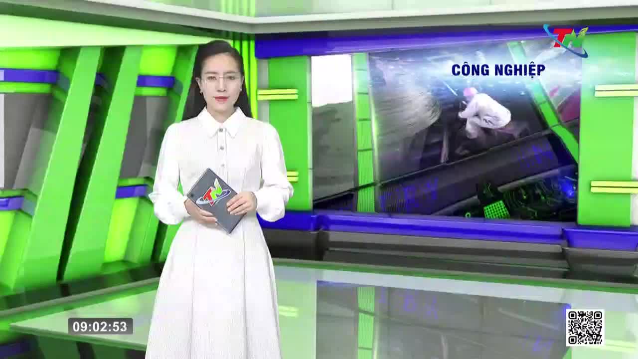 A woman in a white dress stands in a studio, holding a tablet. Behind her, a screen displays a person in protective gear working amidst industrial machinery. The Thai Nguyen TV logo is visible.