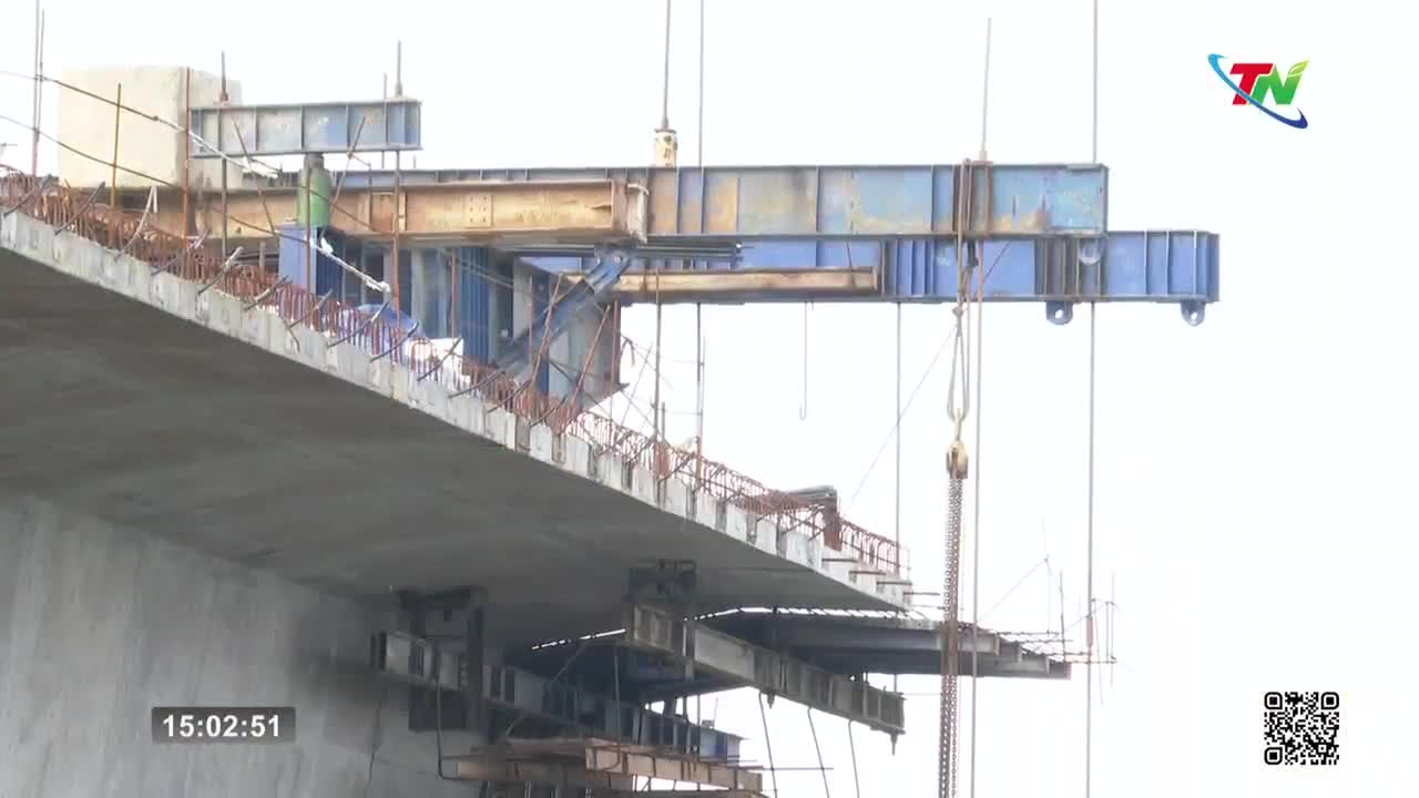 A large crane hoists a section of concrete into place on the bridge under construction. Workers in green vests move around the scaffolding, guiding the massive piece.