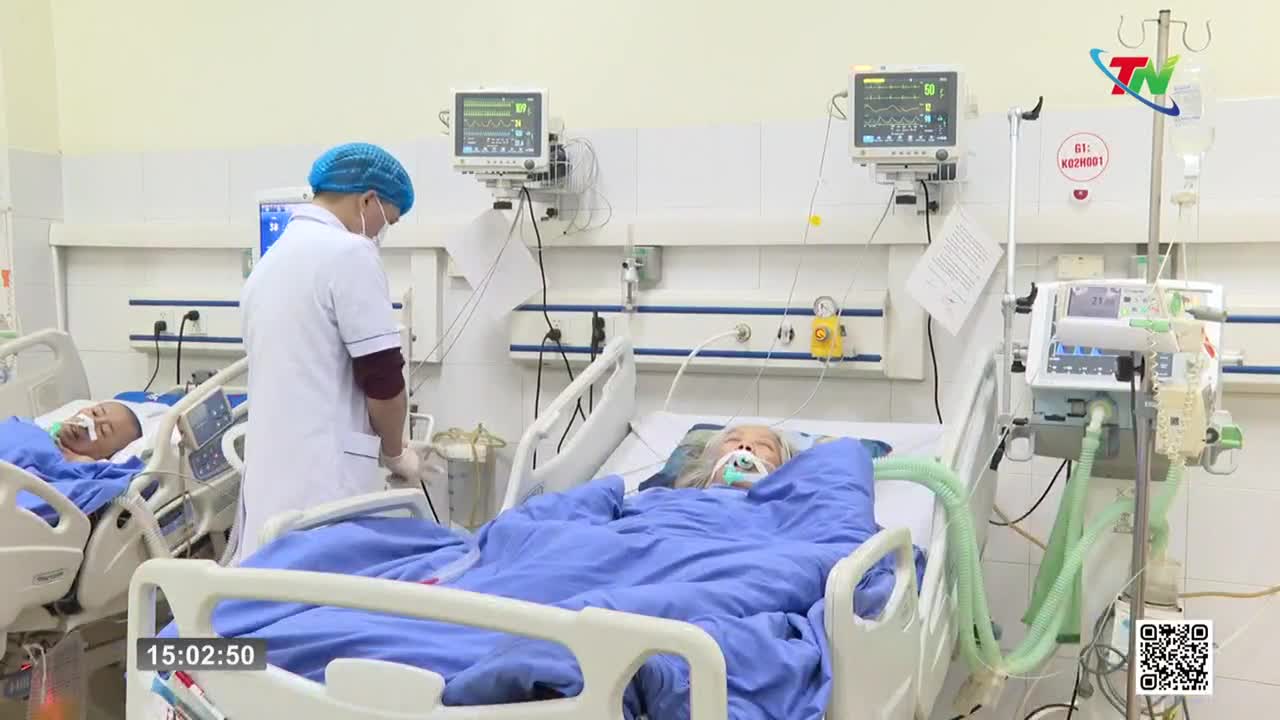 A nurse in a blue cap and mask stands by a patient in a hospital bed, adjusting something near their head. Another patient lies in a nearby bed, connected to a ventilator.