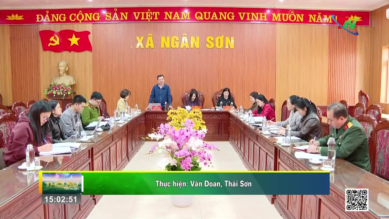 A man in a blue shirt stands at the head of a long table, addressing a group of people seated around it. The room is decorated with Vietnamese flags and a bust of Ho Chi Minh.