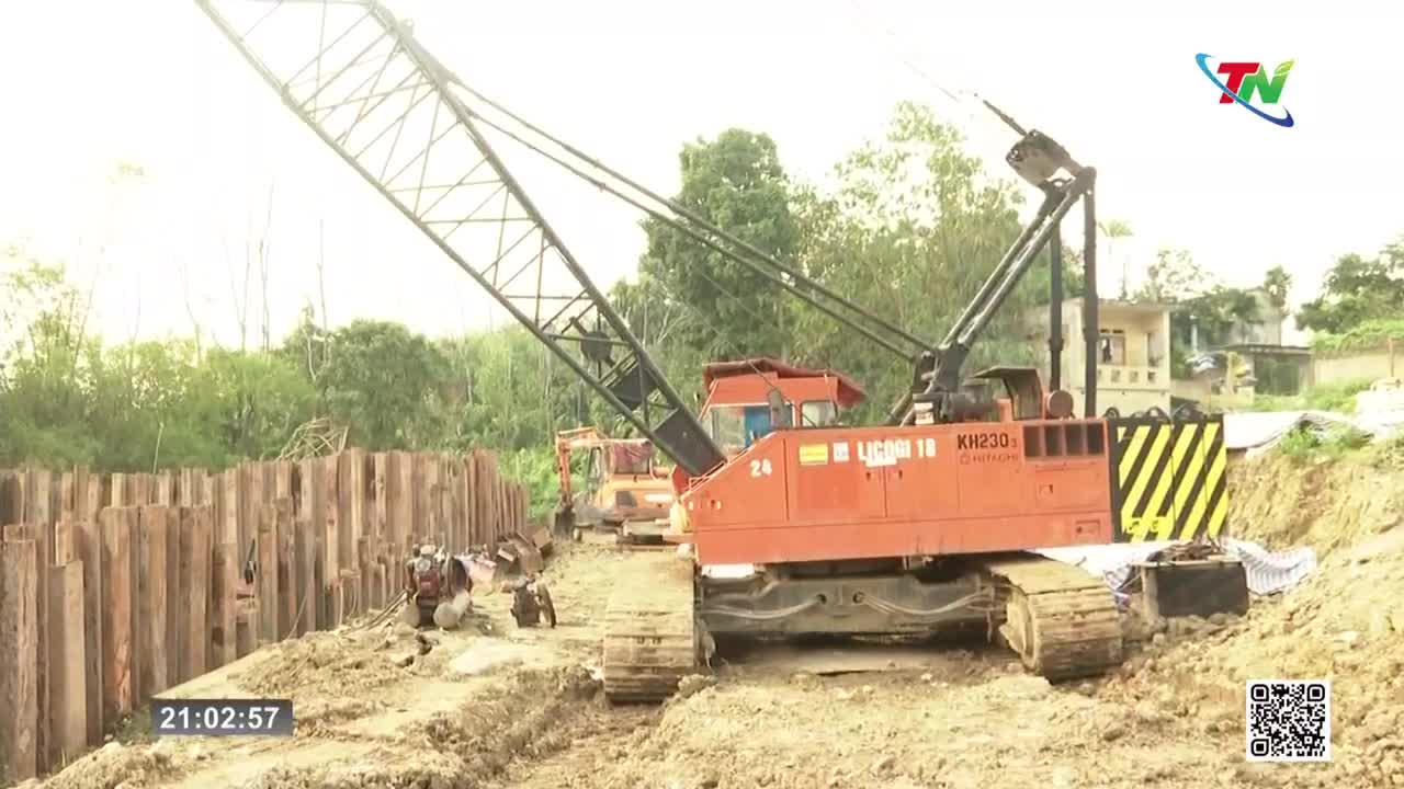 A massive orange crane dominates the scene, its long boom reaching skyward. Workers are busy along a newly constructed wooden retaining wall, the ground churned up around them.