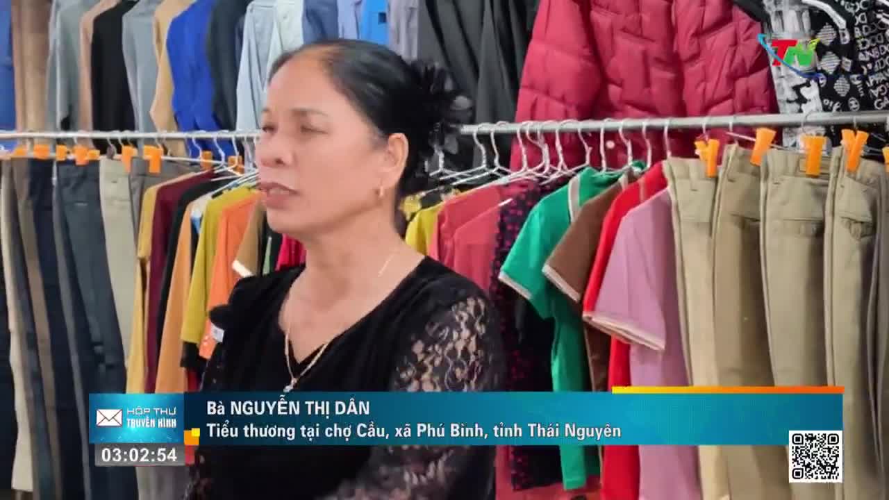 A woman stands in front of racks of clothing, her gaze directed slightly off-camera as if speaking to someone. The garments, mostly shirts and pants, hang neatly on display, a colorful assortment against a backdrop that suggests a bustling market in Thái Nguyên.