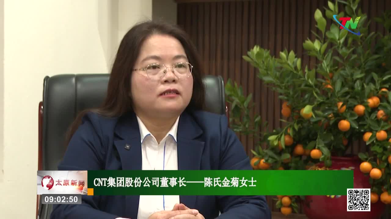 A woman in a dark suit speaks, her hands resting on her lap. Behind her, a small tree is laden with bright orange kumquats.