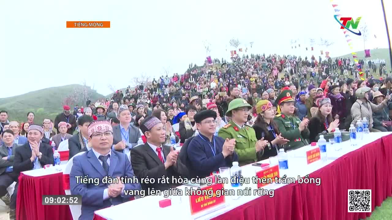 A crowd gathers on a hillside, many wearing traditional headbands. Dignitaries in suits and military uniforms sit at a long table draped in red.