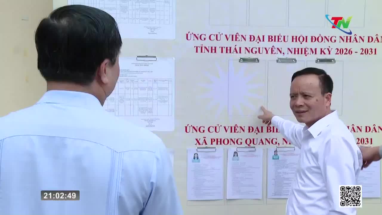 A man in a white shirt points to a bulletin board with candidate information for the Thai Nguyen People's Council. Another man, facing away, stands beside him, looking at the same display.