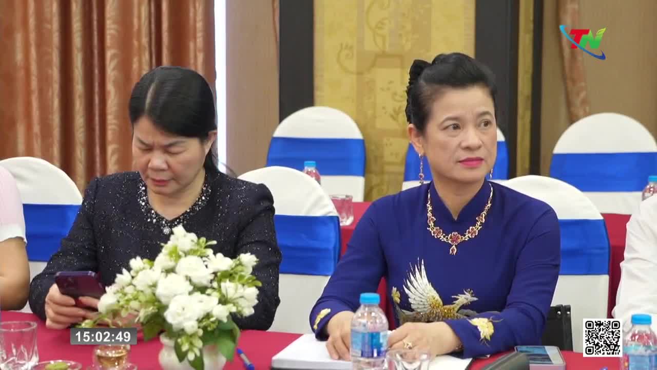 The woman in the blue dress looks attentively to her right, her hand resting on the table. Beside her, another woman glances down at her phone, a bouquet of white flowers between them.