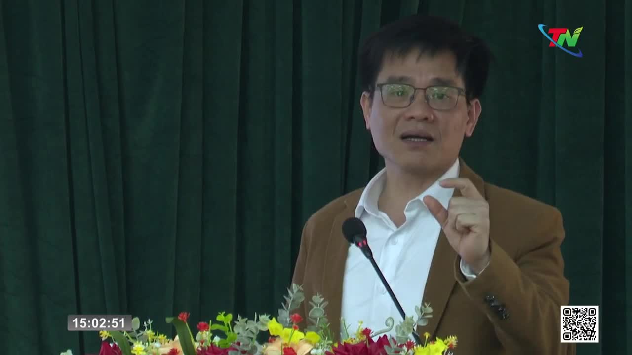 A man in a brown jacket and glasses speaks at a podium, gesturing with his right hand. A microphone stands before him, and a bouquet of flowers rests at the base of the podium.