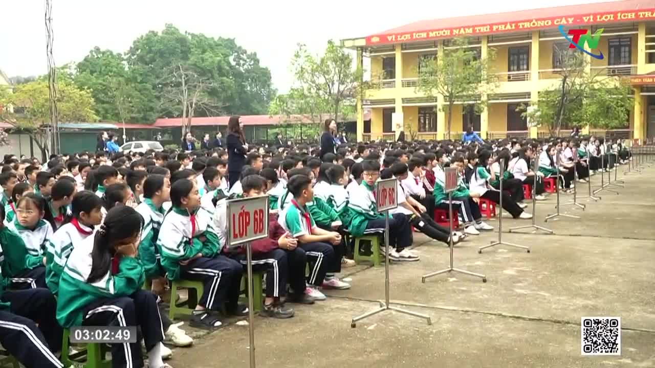Students in green and white uniforms sit in neat rows on a schoolyard in Vietnam. A few adults stand near the front, looking out at the assembled children.