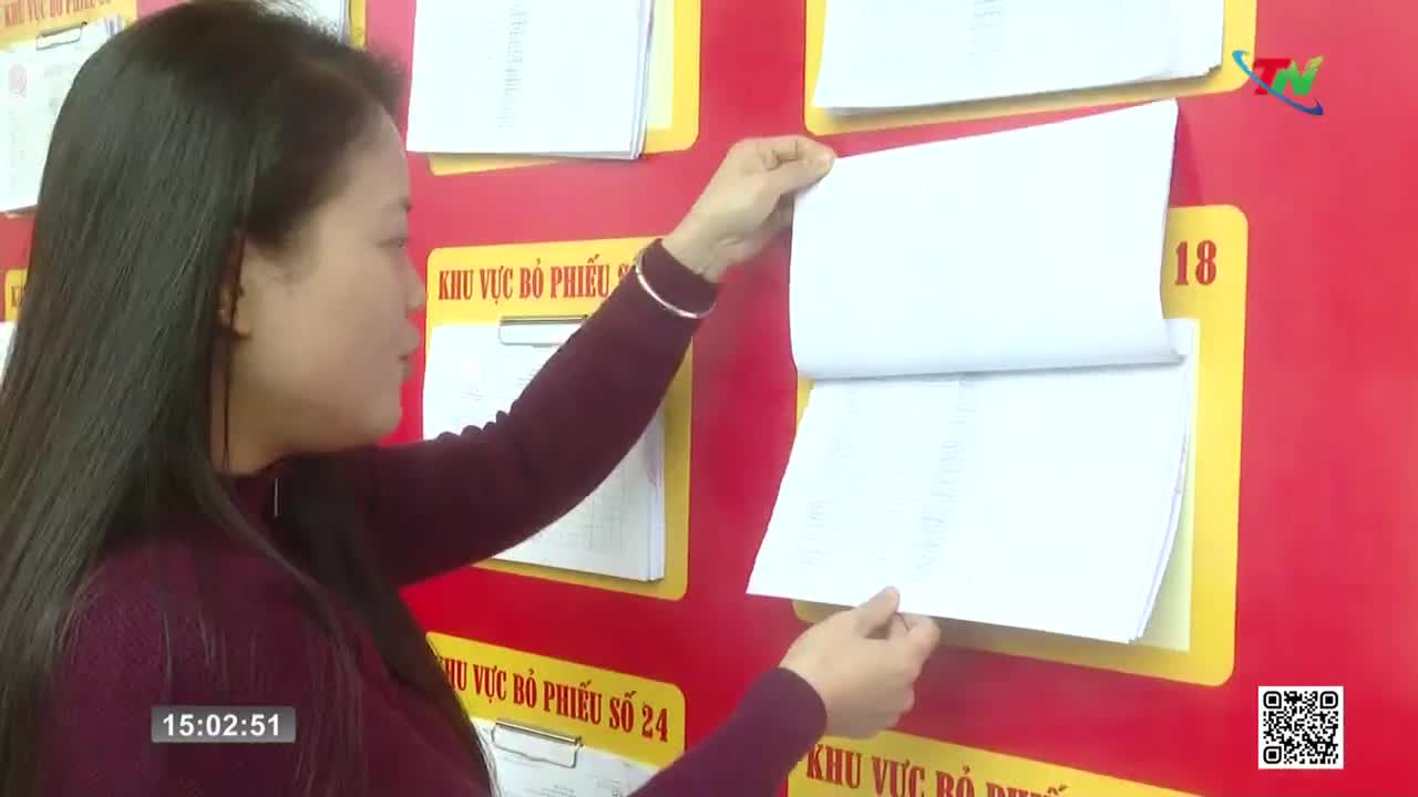 A woman in a maroon sweater adjusts a stack of papers pinned to a bright red wall. The papers are held by yellow clipboards, and some have Vietnamese text.