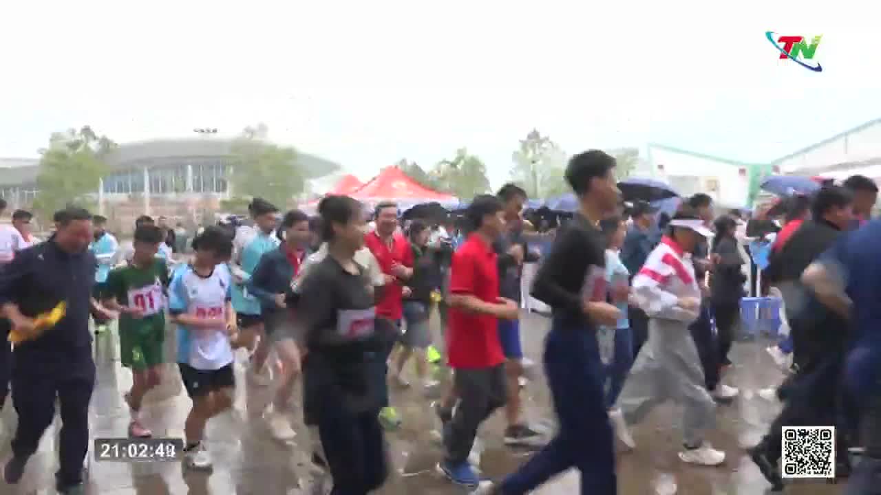 A crowd of runners, many in athletic wear, moves forward on a wet surface. Some hold papers, and a few carry umbrellas as they participate in what looks like a race in Vietnam.