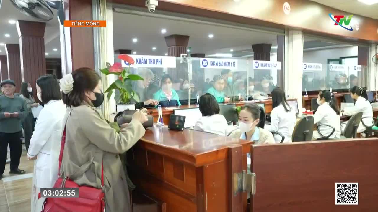 People are lining up at a counter in a busy clinic, likely in Thai Nguyen. A woman in a tan coat is reaching for something on the counter while another woman in a white coat behind the counter looks on.