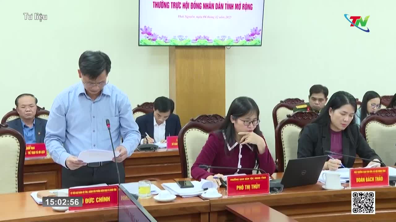 A man in a light blue shirt stands, holding papers and speaking into a microphone. Women seated at a table in front of him are taking notes.