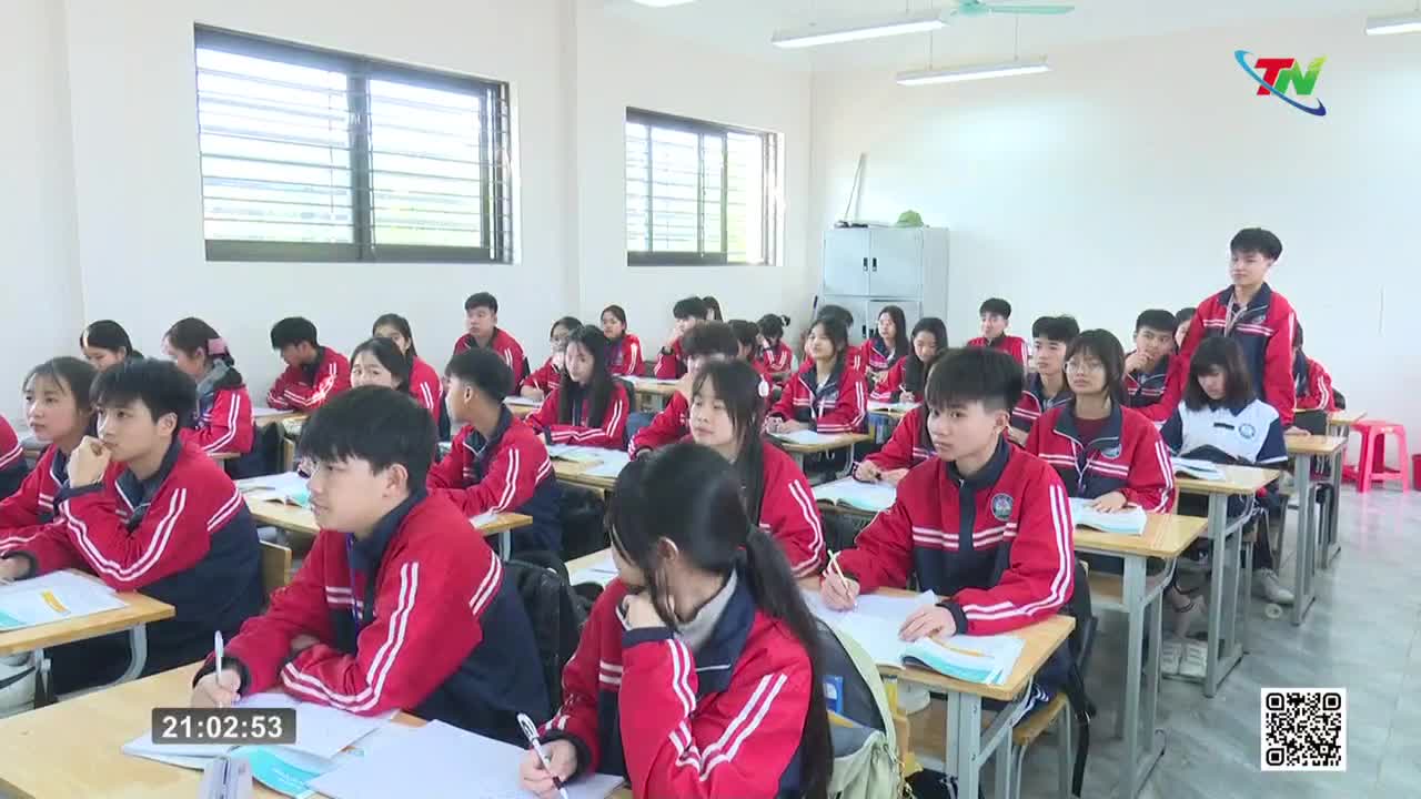 Students in matching red and blue jackets sit at desks, some writing, others looking forward. A young man stands at the back of the classroom, his gaze directed toward the front.