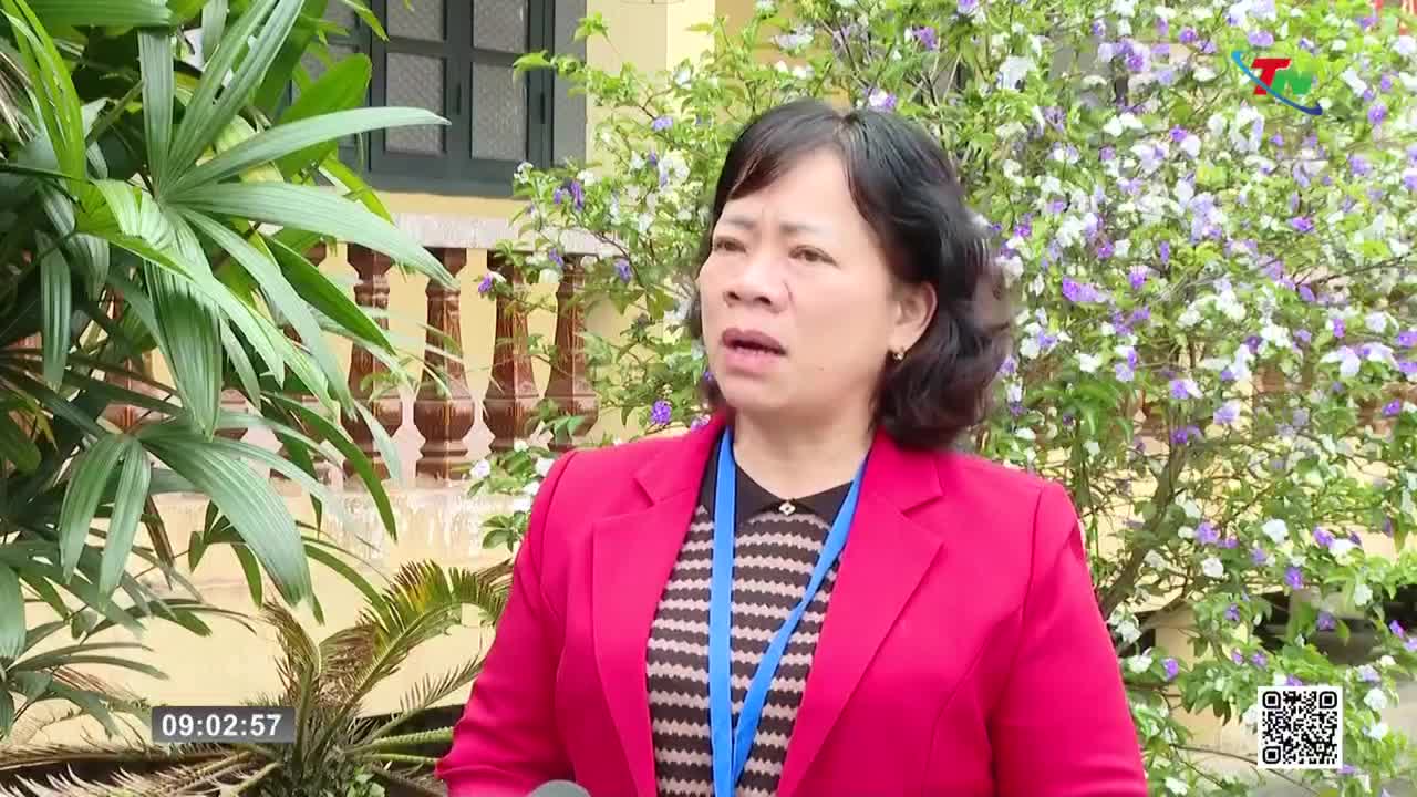 A woman in a bright red jacket speaks into a microphone, her expression earnest. Behind her, a flowering bush bursts with delicate purple and white blooms.