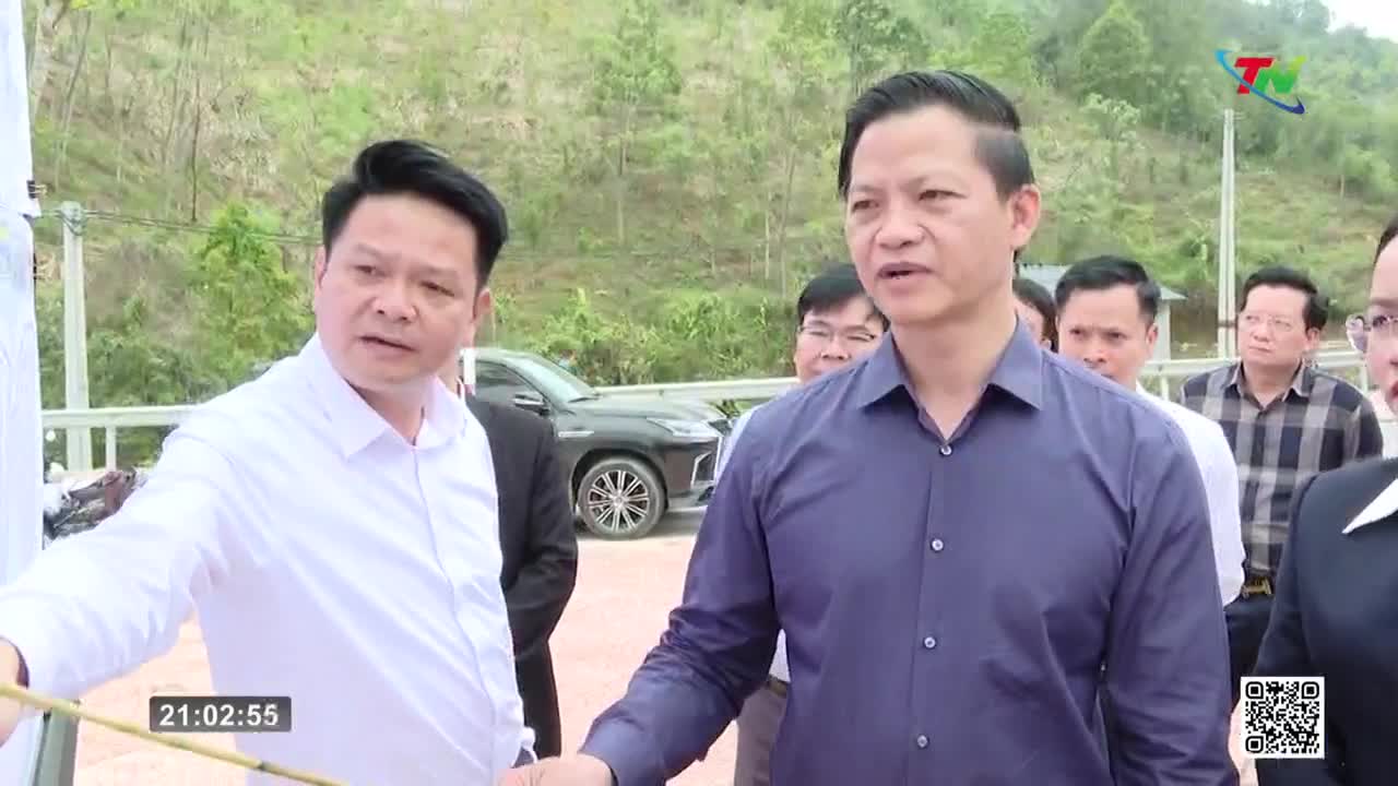 A man in a white shirt gestures towards something off-camera, while a man in a dark blue shirt speaks, his gaze directed forward. Behind them, a black SUV is parked on a road, with a green, hilly landscape in the background.