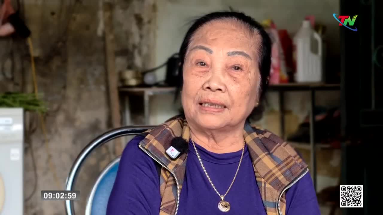 An older woman with dark hair and a patterned vest speaks directly to the camera. A small microphone is clipped to her vest, and a pendant hangs from a necklace.