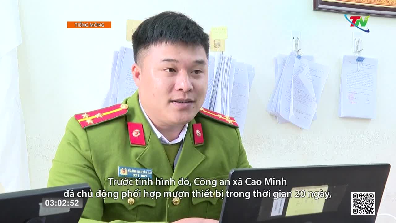 A police officer in a green uniform speaks at a desk. Papers are stacked behind him on the wall.