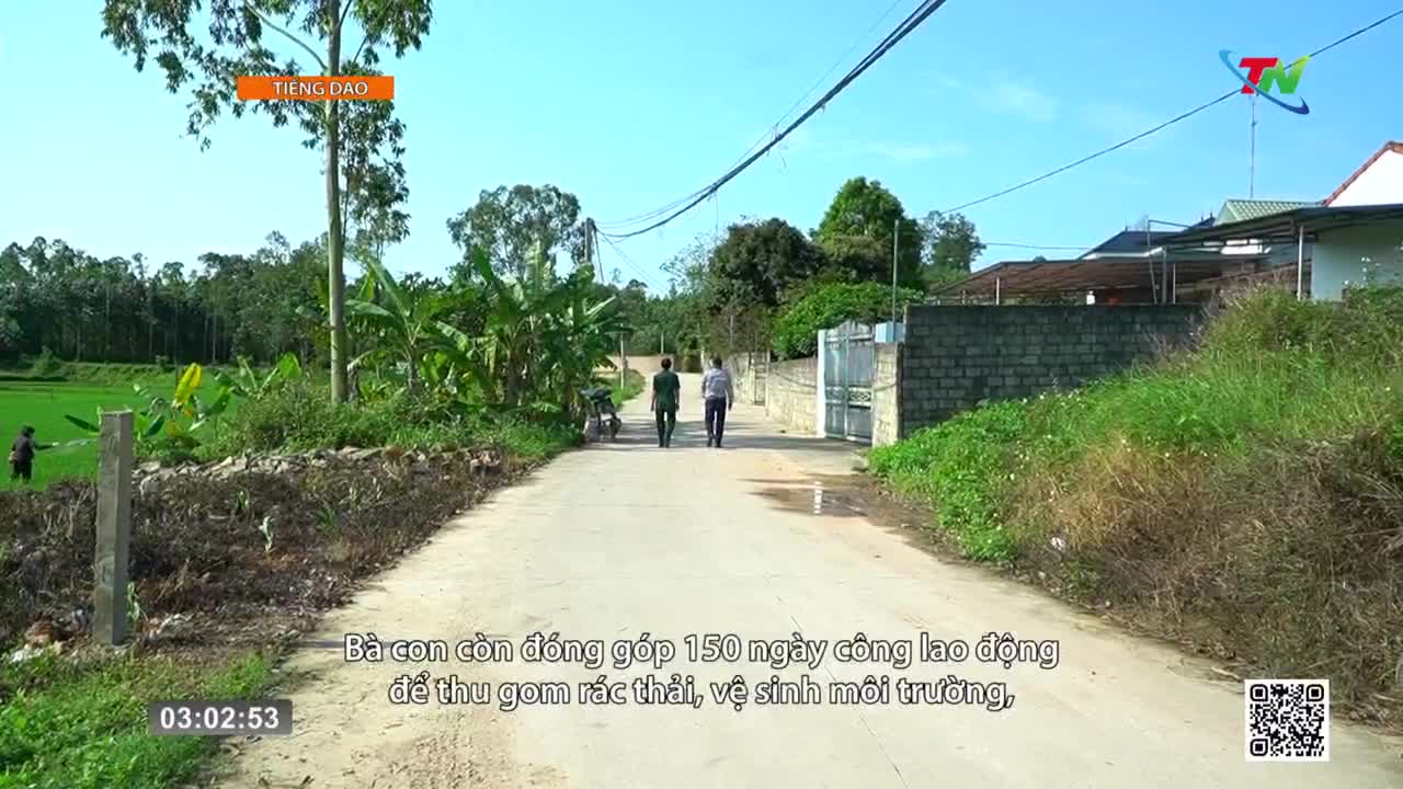 Two men walk down a rural road in Vietnam, the sun bright overhead. A person works in a green rice paddy to the left, and a motorbike is parked by the side of the road.