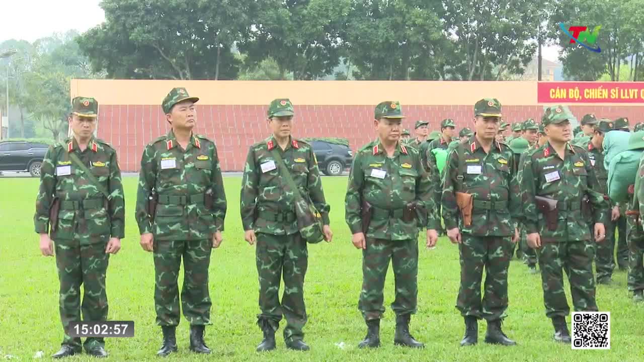 A line of soldiers in camouflage stands at attention on a grassy field. Behind them, more soldiers are gathered, and a banner with Vietnamese text hangs on a wall.